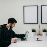 Young man in a hoodie works on a laptop in a modern, minimalist home office setup.