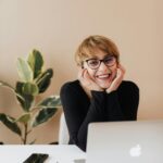 Cheerful woman in eyeglasses and black sweater smiling while working at a desk with a laptop indoors.